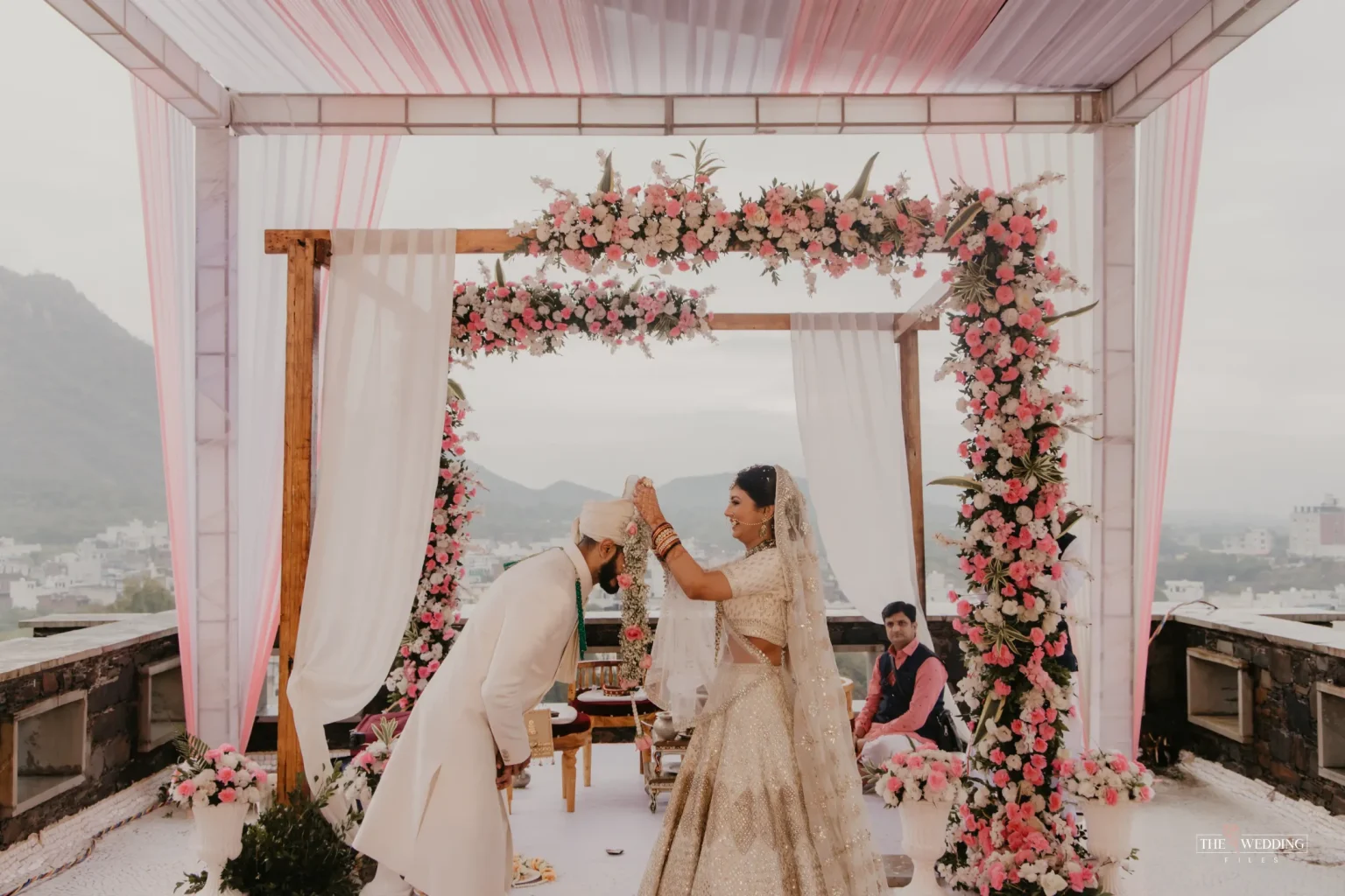 Bride in ivory gold lehenga and groom in white sherwani exchanging wedding vows under a wooden mandap decorated with pink and white flowers, with Udaipur hills and cityscape in the background during a rooftop destination wedding ceremony