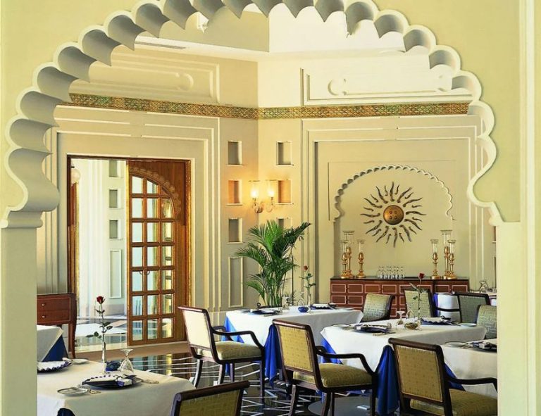 Grand restaurant interior at The Oberoi Udaivilas, featuring ornate arches, a decorative ceiling, chandeliers and patterned marble flooring.