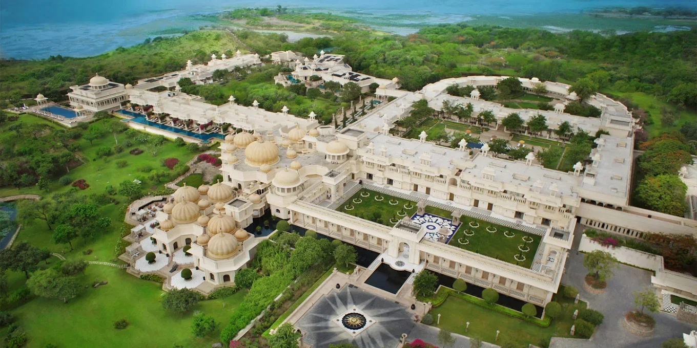 Panoramic aerial view of The Oberoi Udaivilas palace resort in Udaipur, with landscaped gardens, domed buildings and swimming pools.
