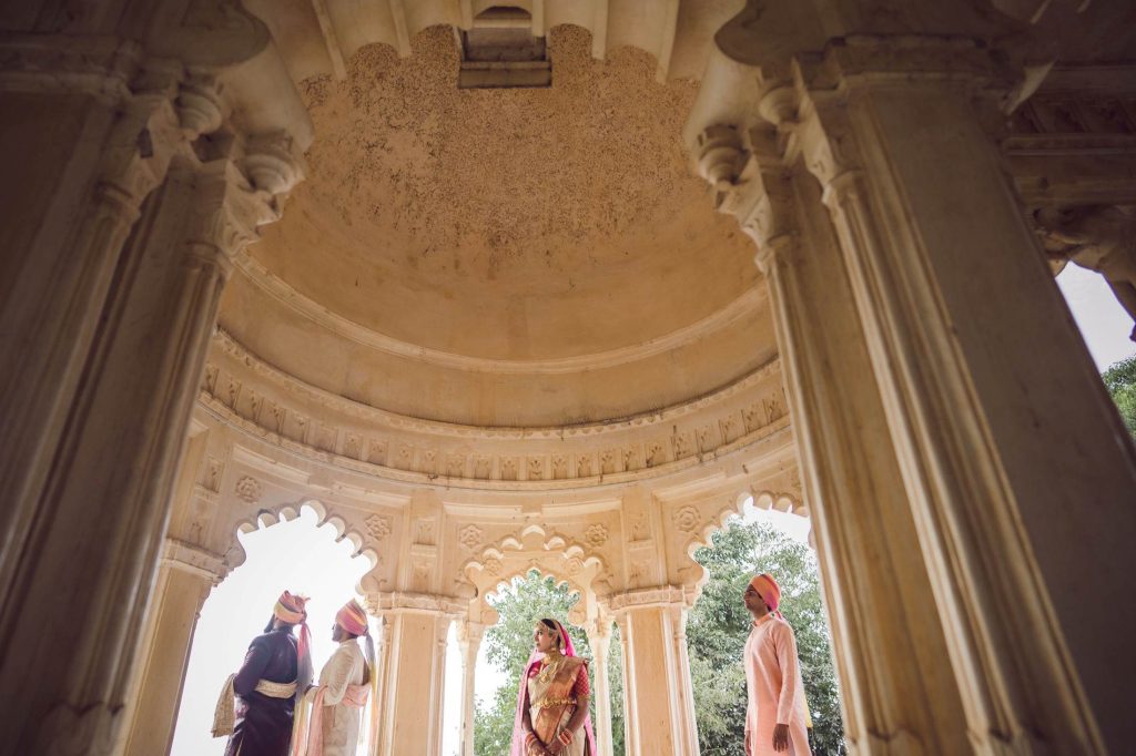 Bride in pink bridal saree with gold jewellery standing inside ornate carved marble chhatri pavilion with Rajasthani musicians at heritage palace wedding