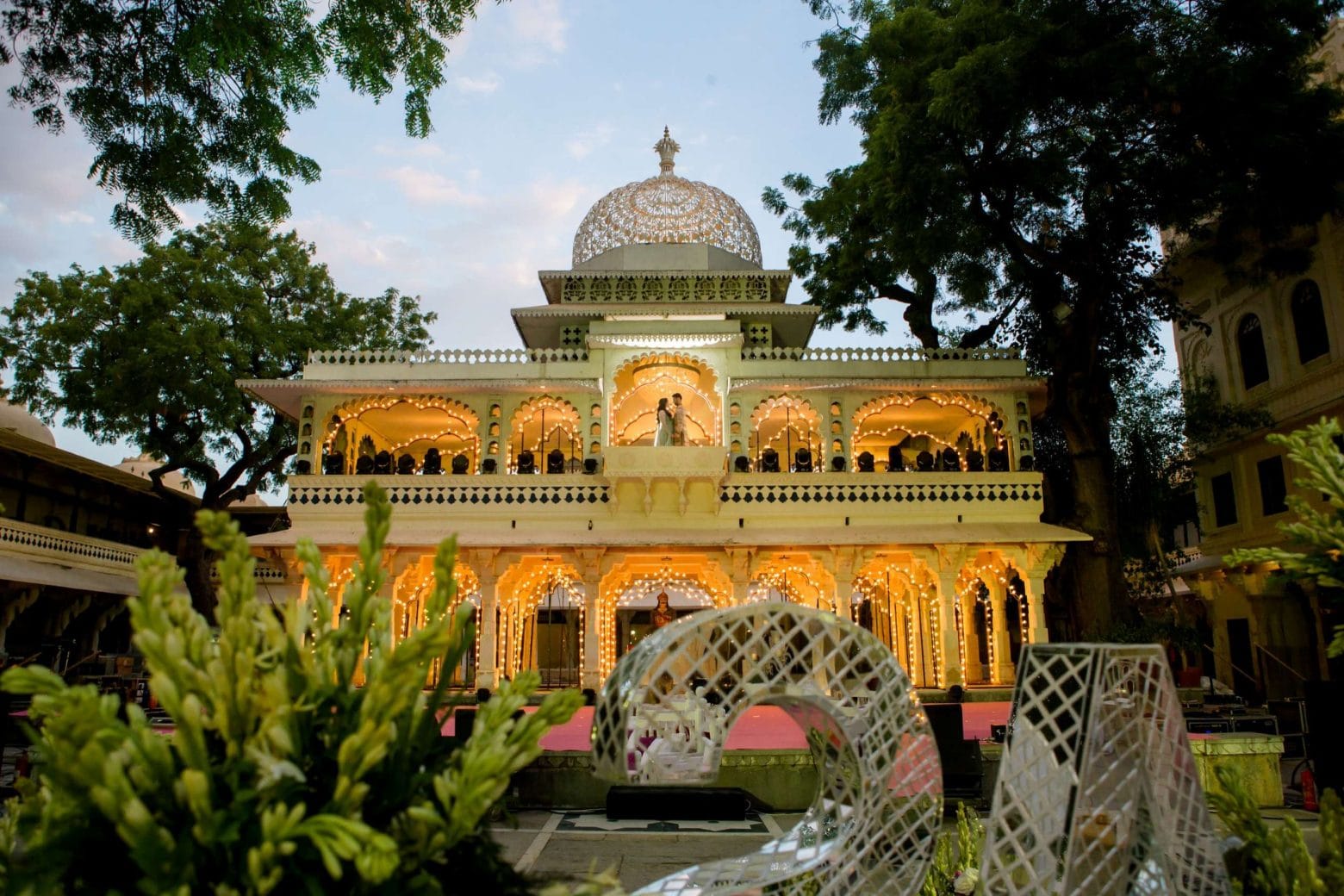 Illuminated multi-storey Rajasthani palace at dusk with couple visible on upper balcony and wedding decor setup in foreground at destination wedding in Udaipur