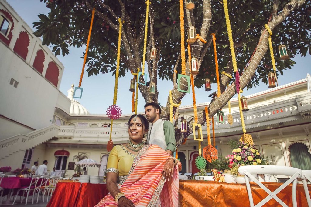 Bride in orange striped saree and groom in green kurta posing under tree decorated with colourful hanging lanterns and marigold garlands at Rajasthani palace courtyard during mehendi