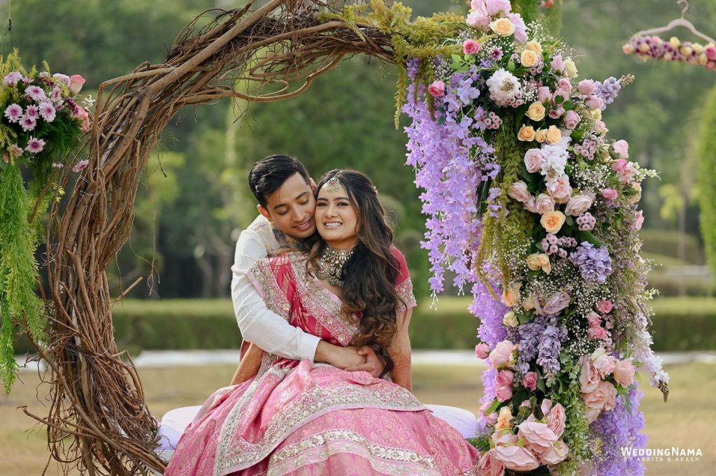 Couple posing under rustic wooden arch with cascading pink and lilac flowers at outdoor engagement shoot