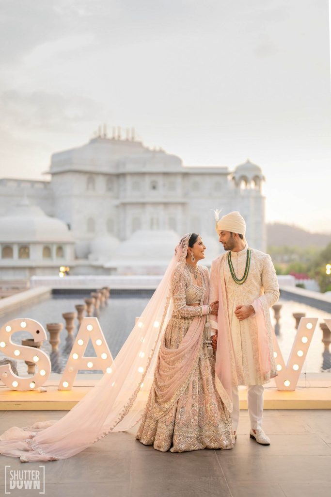 Bride in blush lehenga and groom in cream sherwani with illuminated monogram letters at Udaipur palace wedding