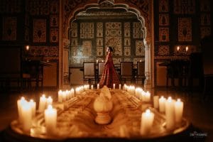 Bride in red lehenga standing inside ornate mirror work hall framed by rows of pillar candles at The Leela Palace Jaipur, luxury destination wedding by Desert Pearl Weddings