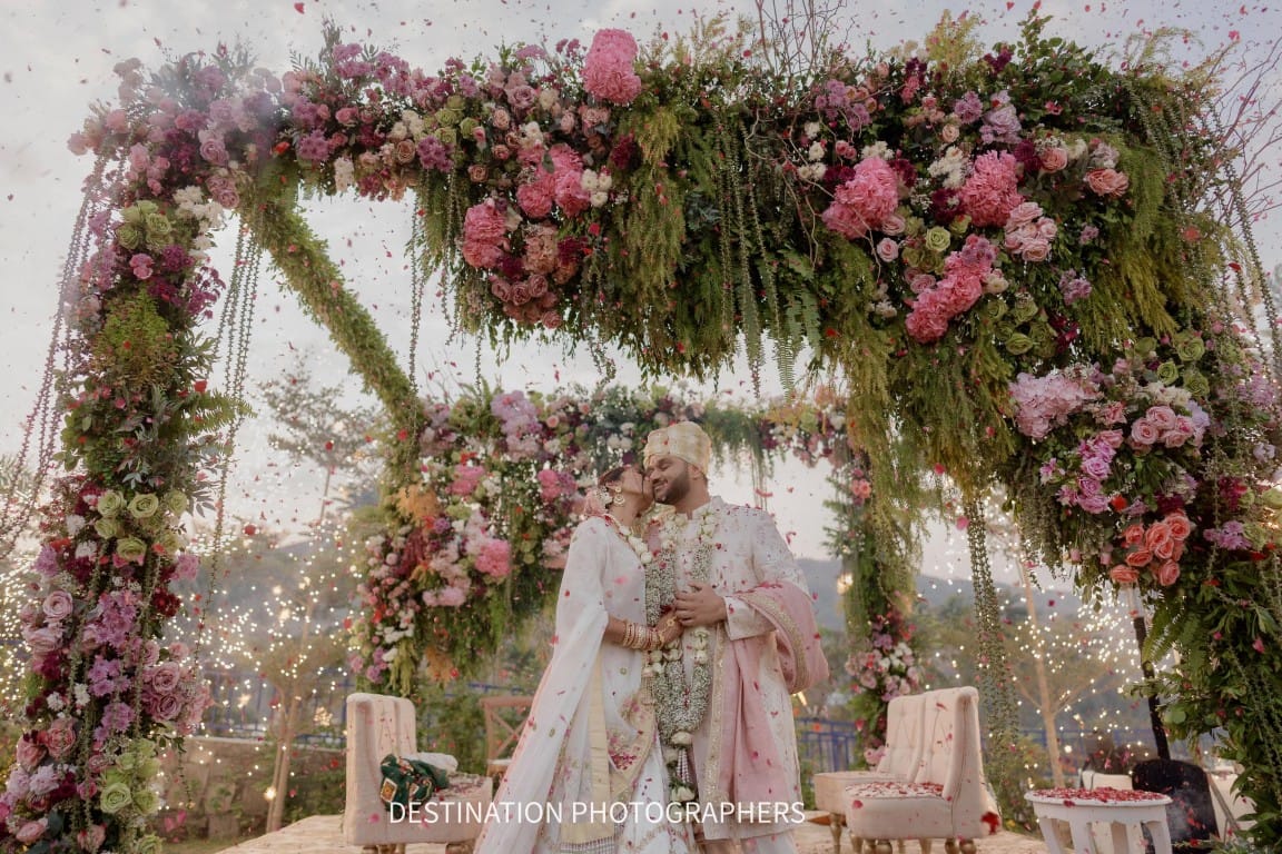 Bride and groom under a grand pink and white floral mandap arch with rose petals falling at a luxury destination wedding in Udaipur, Rajasthan — Desert Pearl Weddings