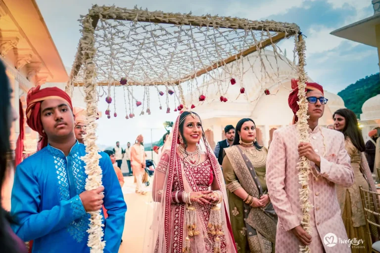 Bride in a deep crimson red heavily embroidered lehenga with a sheer blush pink dupatta draped over her head wearing a layered multi-strand gold and pearl necklace set large gold jhumka earrings a gold maang tikka and red and white chooda bangles with long gold tassel kalire hanging from her wrists walking with a composed slightly smiling expression under a large square flat floral chadar canopy densely covered in white jasmine flowers with small red rose buds hanging from the edges held aloft on four poles by family members including a young man in a bright cobalt blue kurta with a red turban on the left and a young man in a pale blush pink embroidered sherwani with a dark red turban and blue-rimmed glasses on the right with a woman in an olive gold embroidered saree walking beside the bride and a white heritage palace dome and Aravalli hills visible under a dusk blue sky in the background at a luxury destination wedding in Rajasthan