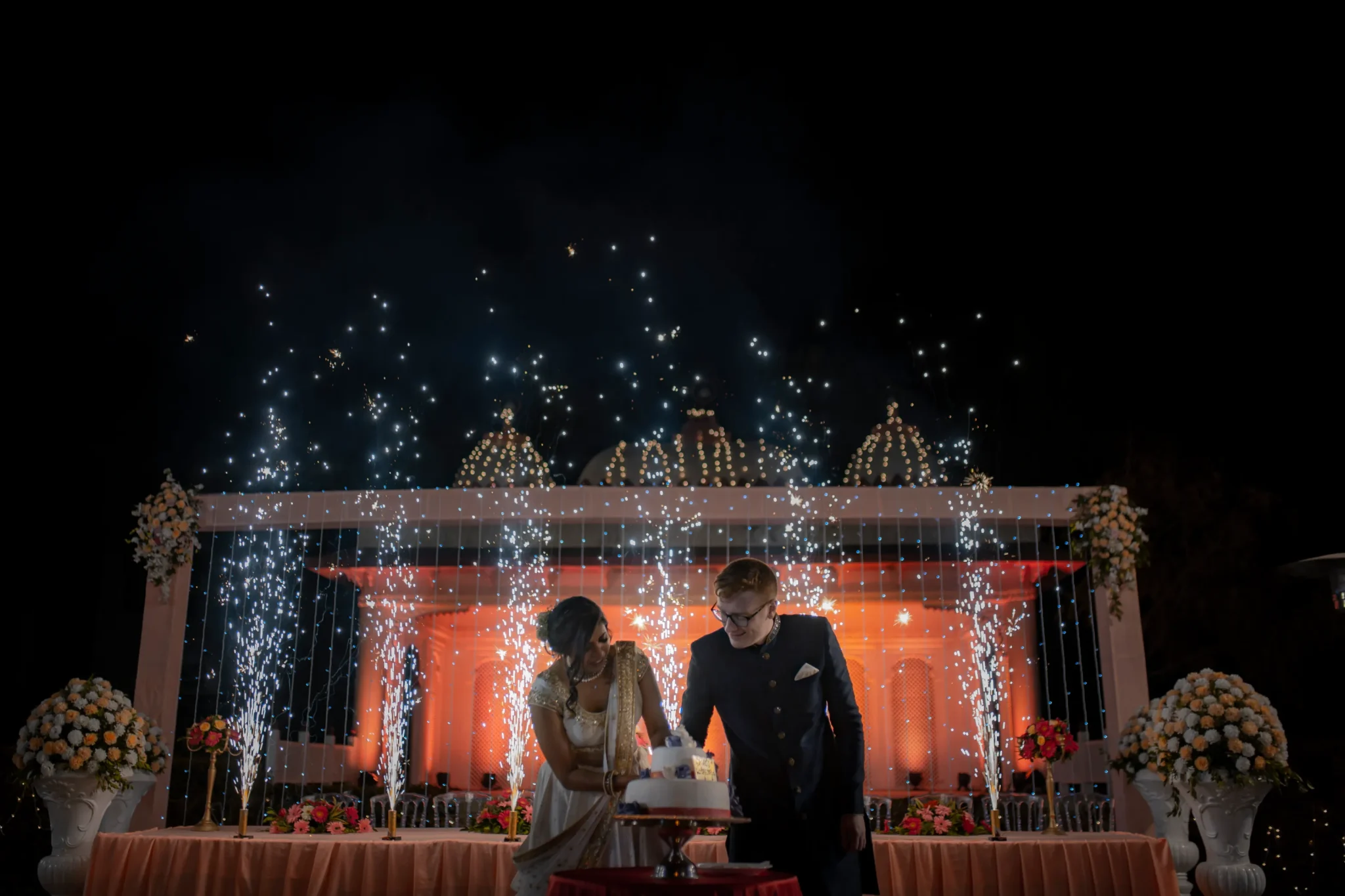 Couple bending together over a white fondant wedding cake decorated with a small cake topper on a table draped in pale blush linen with the bride in a gold champagne embroidered saree leaning in and the red-haired groom in a dark navy sherwani with glasses standing beside her as a dramatic silver cascading sparkler waterfall curtain falls behind them and starburst cold pyrotechnics shoot upward above a large decorative lattice white stage backdrop illuminated in deep warm orange with tall white urn floral arrangements on both sides of the cake table against a completely dark night sky at a luxury destination wedding reception in Rajasthan