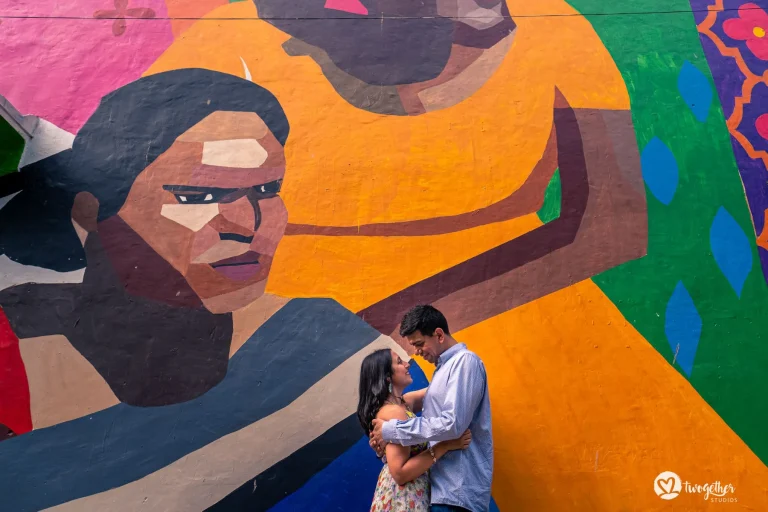 Man in a light blue cotton button-down shirt holding a woman in a bright floral spaghetti-strap dress close as they lean their foreheads together with soft smiles in front of a massive bold colorful street art mural covering the entire wall featuring a large geometric painted face in warm brown and orange tones with bold pink green blue and purple abstract shapes filling the background at a pre-wedding couple shoot in Rajasthan