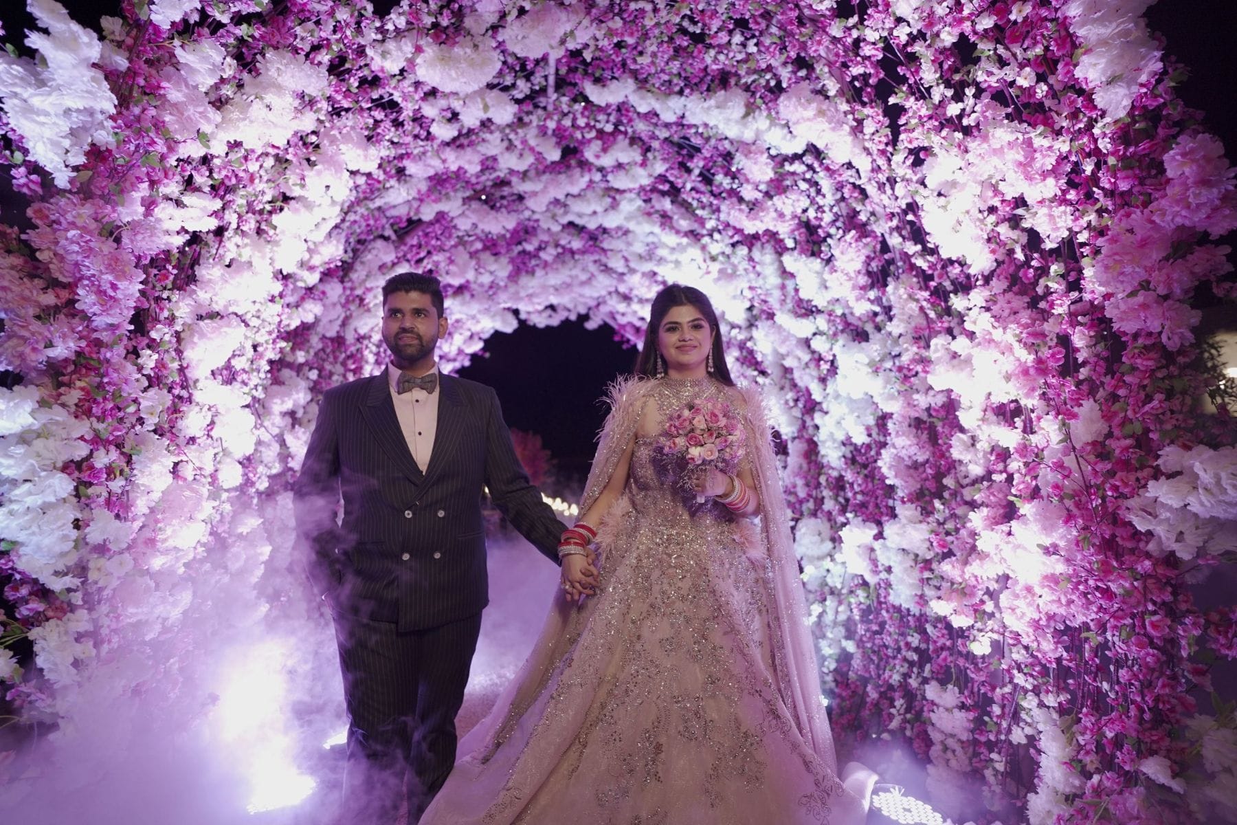 Bride in a heavily embroidered silver and blush ball gown lehenga with feathered cape holding a pink rose and lavender bouquet and groom in a dark pinstripe suit with bow tie walking hand in hand through a dramatic arched tunnel of pink and white flowers with dry ice fog swirling around their feet at a luxury destination wedding night celebration in Rajasthan