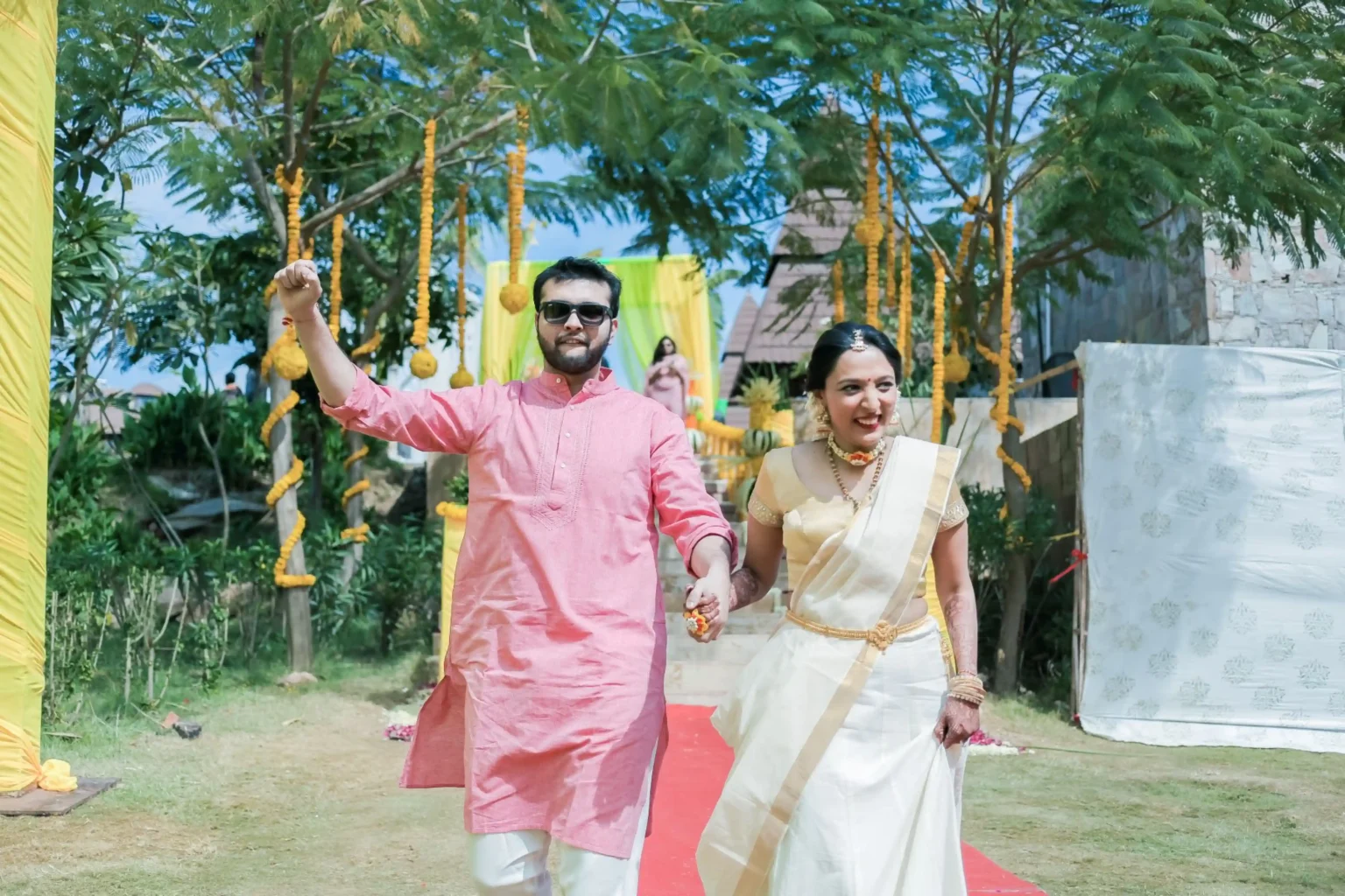 Groom in pink kurta and white mundu with sunglasses raising his fist in the air while holding bride's hand as she walks in a traditional cream Kerala kasavu saree with gold border and gold jewellery along a red carpet outdoor pathway decorated with hanging marigold garlands on trees at a destination wedding haldi entry in India