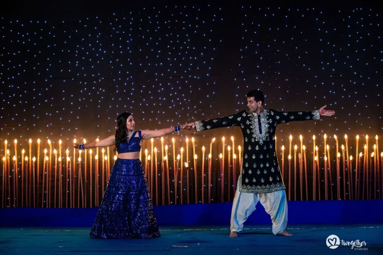 Couple performing a choreographed sangeet dance on a blue-lit stage with both arms extended wide apart holding hands at fingertips with the woman in a royal cobalt blue heavily sequined mirror-work lehenga with a cropped blouse and mehndi-covered hands wearing blue bangles and the man in a deep black embroidered sherwani with gold zari motifs and white dhoti-style pants both barefoot against a dramatic backdrop of a long row of tall burning torch flames lined across the full width of the stage with a dense dark blue LED starry night effect panel above at a luxury destination wedding sangeet in Rajasthan