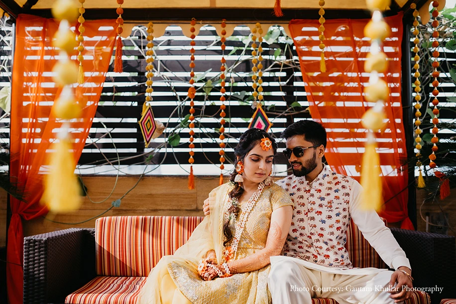 Bride in an ivory cream embroidered lehenga with a side-parted braid adorned with a large orange flower hair accessory and layered white floral gajra garland necklace with mehndi-covered hands resting in her lap gazing downward with a serene expression while the groom beside her in a white kurta printed with small red elephant motifs and dark sunglasses drapes his arm around her as they sit together on a red and white candy-striped cushioned sofa seat under a vibrant orange fabric canopy decorated with hanging orange and yellow bead tassel strings diamond-shaped colorful pendants and a black and white horizontal slatted backdrop with green tropical vines above at a luxury destination wedding mehendi ceremony in Rajasthan