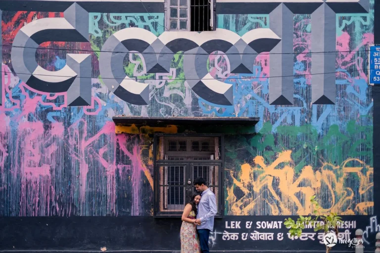 Woman in a light floral dress and man in a pale blue shirt standing close together with the man leaning his head down toward her and both looking downward in a tender quiet pose against a large dark grey building exterior completely covered in layered urban graffiti art featuring large stylized black and white geometric lettering with splashed drips of bright pink yellow green and blue paint and a barred window at center with a blue street number signboard visible at the upper right and small green leaves of a potted plant at the lower edge at a pre-wedding couple shoot in Rajasthan