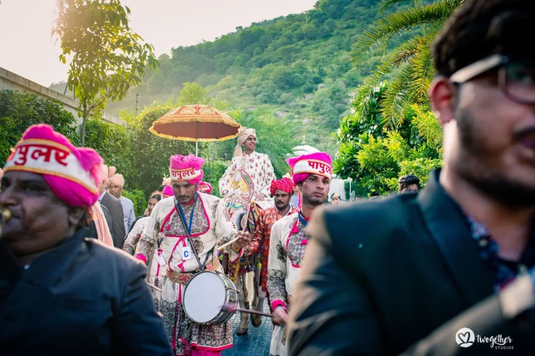 Groom in a cream ivory embroidered sherwani and a peach safa turban seated elevated above the crowd on a decorated ceremonial doli palanquin being carried through a narrow tree-lined path smiling warmly as a traditional wedding band of musicians wearing hot pink turbans with gold and pink embroidered uniforms march ahead playing drums with drumsticks and one musician blowing a wind instrument while guests in formal wear including a man in a dark suit with sunglasses appear in the close foreground with dense lush green Aravalli hills and trees rising steeply in the background in warm golden haze light at a luxury destination wedding baraat in Rajasthan