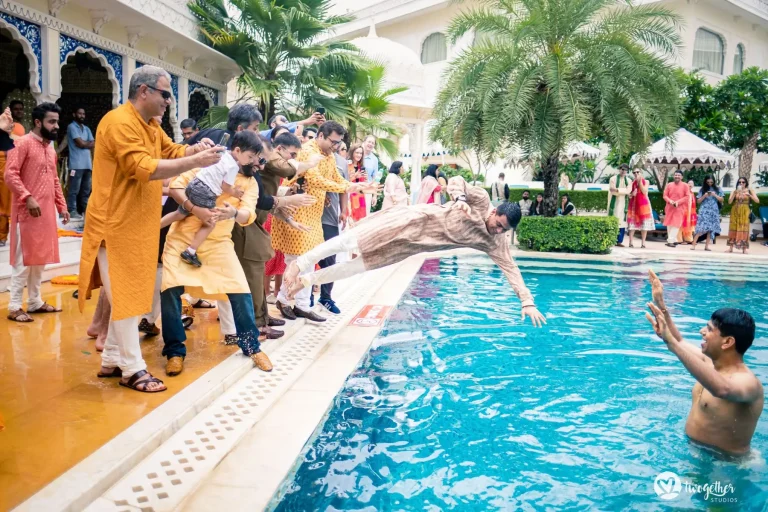 Groom in a beige textured kurta and white churidar mid-dive in a horizontal position launching himself headfirst from the white marble pool edge into a bright turquoise blue swimming pool as a shirtless male guest already in the water raises his arms to catch him while a large group of guests in colorful ethnic wear including mustard orange pink and red kurtas and salwars line the pool edge leaning forward cheering photographing and pointing from the side with a grand white heritage palace building featuring blue and white ornate tile and arch detailing palm trees and tropical foliage visible in the background at a luxury destination wedding haldi celebration in Rajasthan