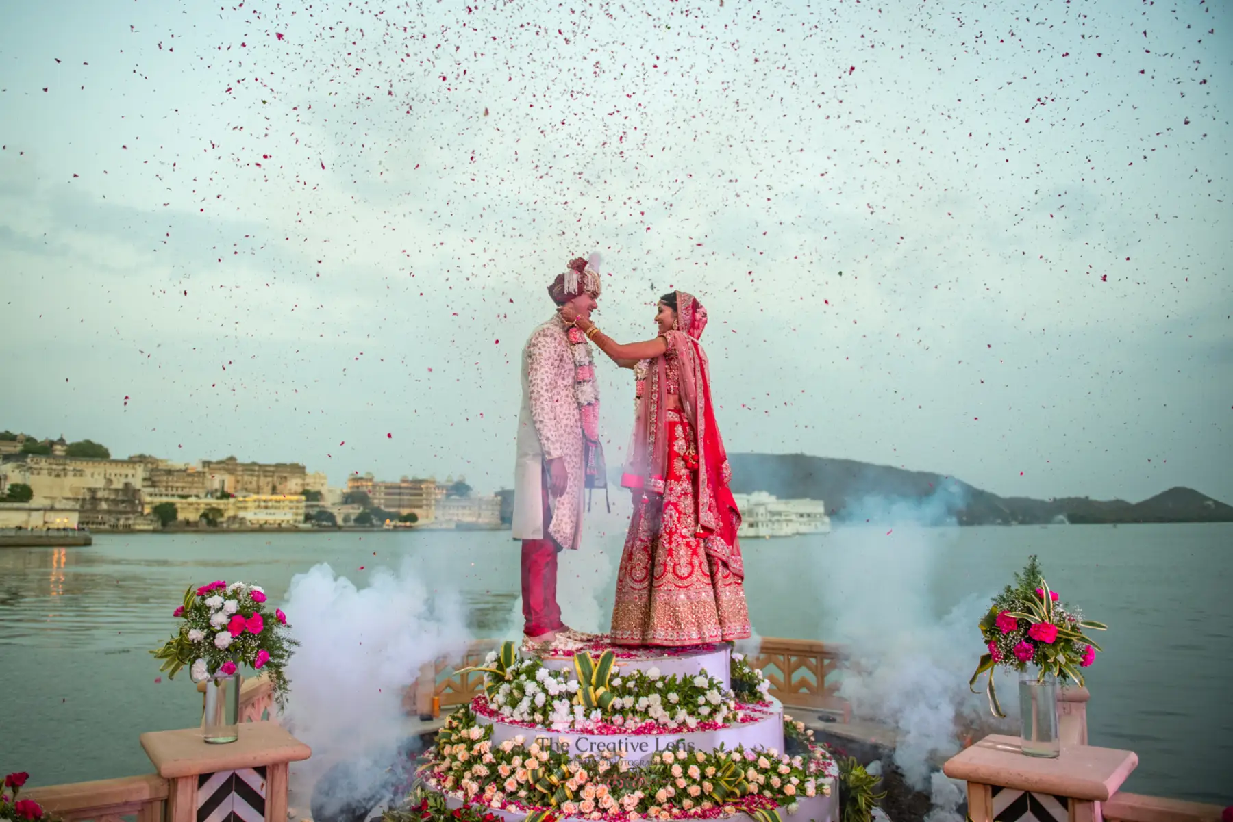 Bride in a red embroidered bridal lehenga and groom in a white floral sherwani with pink dupatta exchanging garlands on an elevated floating stage surrounded by dry ice smoke and a dramatic shower of red and white rose petals with Lake Pichola and the City Palace of Udaipur visible in the background at a luxury destination wedding