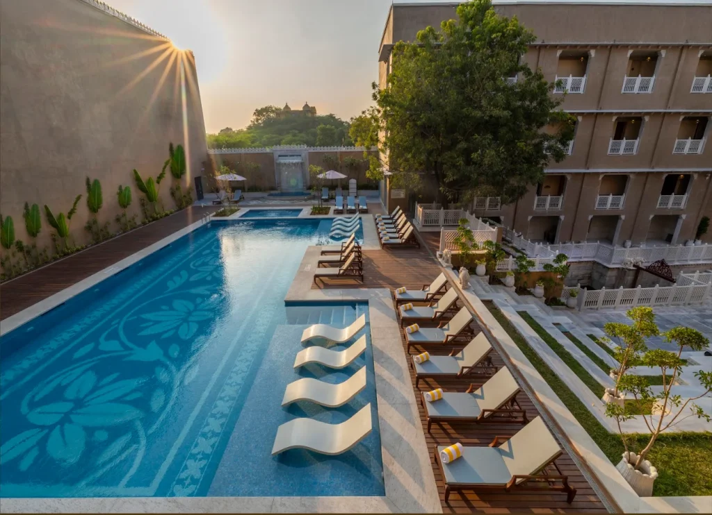 Aerial pool terrace with wave sun loungers at golden hour and Monsoon Palace silhouette at Taj Lalit Bagh Udaipur