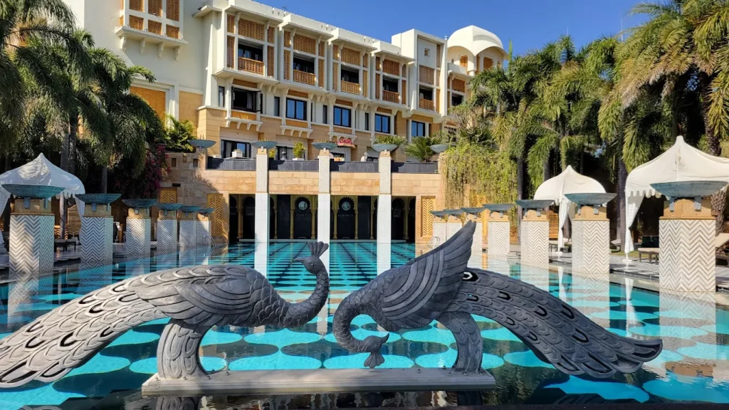 Black peacock sculptures flanking teal geometric pool with chevron columns and palm backdrop at Trident Udaipur