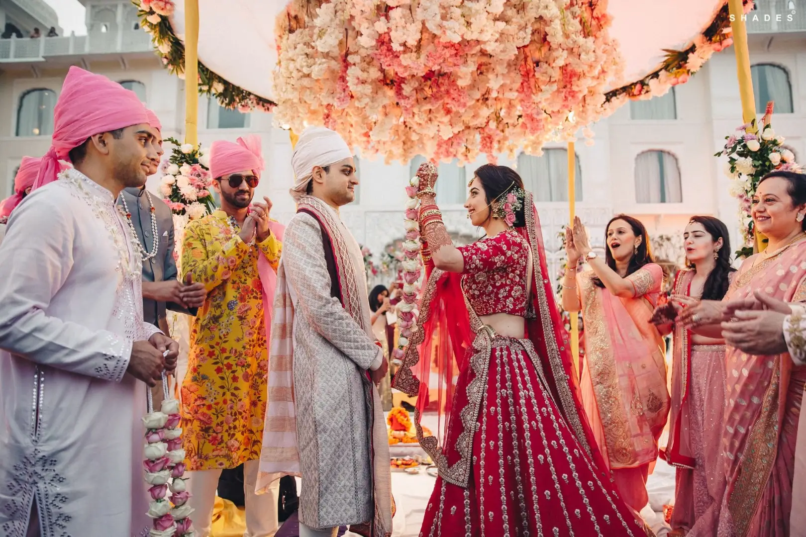 Bride in a heavily embroidered red bridal lehenga raising a pink and white rose garland toward groom in a cream brocade sherwani with white safa turban as groomsmen in pink turbans and yellow floral kurtas watch laughing from the left while female family members in peach and pink sarees clap with joy on the right beneath a large circular pink and cream floral chandelier with a white heritage palace building visible in the background at an outdoor wedding in Rajasthan