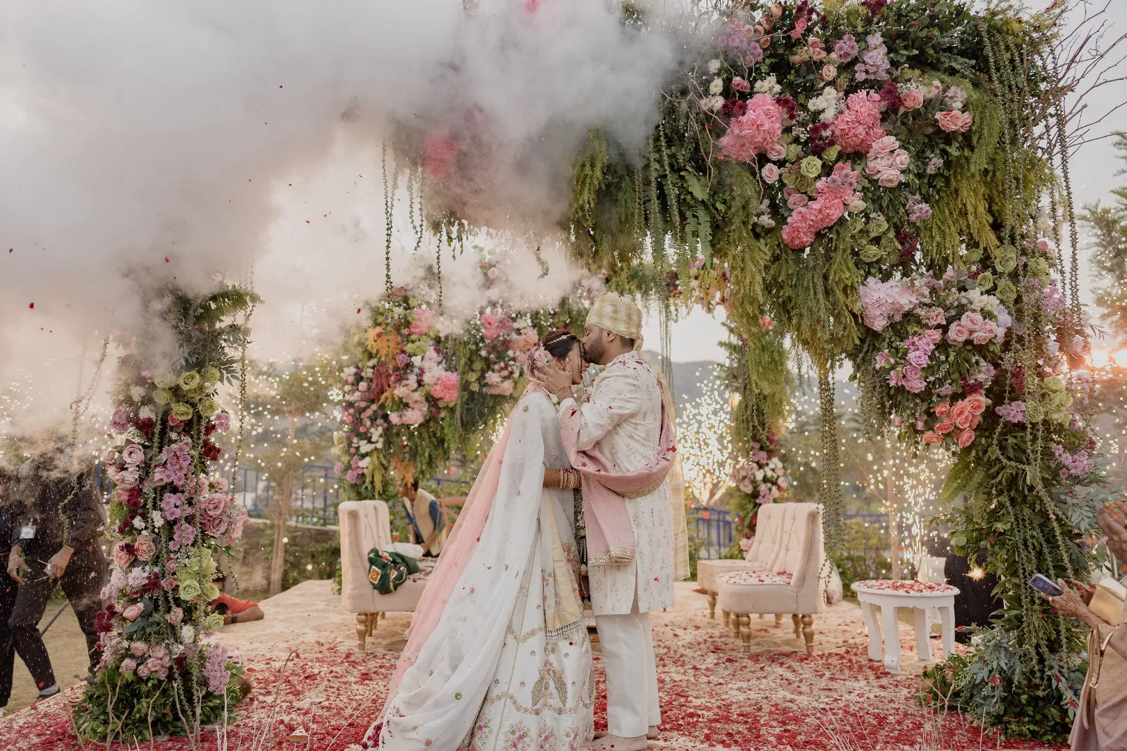 Bride and groom sharing a close romantic moment at the conclusion of their wedding ceremony surrounded by sparklers and pink smoke under an elaborate outdoor arch decorated with pink roses, hydrangeas, and cascading green foliage at a luxury destination wedding in Rajasthan