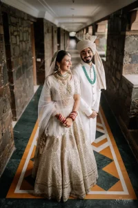 Bride in white net lehenga with green polki bridal jewellery and groom in ivory sherwani with cream turban and turquoise mala standing together in a heritage hotel corridor at a luxury destination wedding in Udaipur Rajasthan