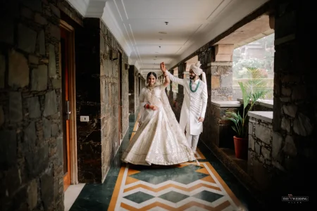 Bride in full white sequined lehenga spinning joyfully while groom in white sherwani and turban holds her hand raised, captured in a geometric-floored corridor at a luxury heritage hotel in Udaipur on their wedding day