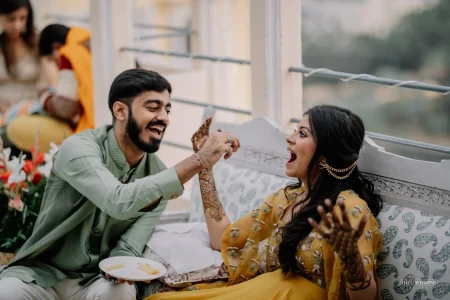 Couple sharing a candid joyful moment during mehendi ceremony, groom in mint green sherwani laughing while feeding something to bride in mustard yellow anarkali with intricate mehndi on her hands, with flowers in background