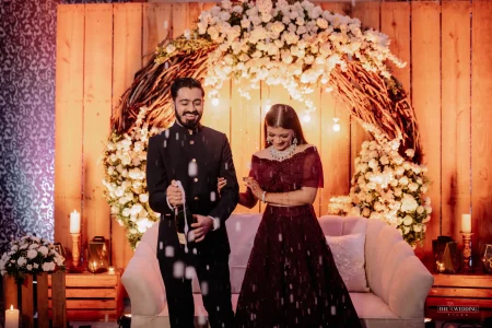 Couple popping a champagne bottle laughing together at their engagement ceremony with a white floral circular arch backdrop and candlelit wooden stage, groom in dark navy bandhgala and bride in maroon off-shoulder lehenga at a luxury Udaipur wedding venue