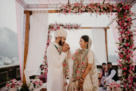 Groom in white sherwani gently kissing bride's hand while she laughs during their wedding ceremony under a wooden mandap decorated with pink and white flowers, misty Aravalli hills visible in the background at an outdoor Udaipur wedding