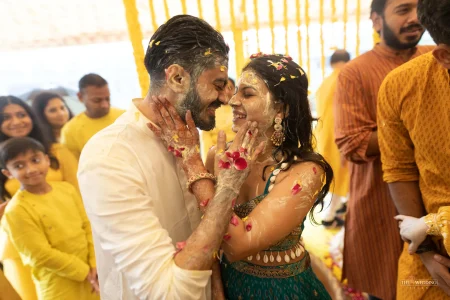 Couple sharing an intimate laughing moment during haldi ceremony with turmeric paste on their faces and rose petals scattered around, surrounded by guests dressed in yellow at a destination wedding in Udaipur