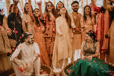 Couple seated during haldi ceremony being showered with red rose petals by smiling friends and family, bride in teal lehenga and groom in white kurta, at a multi-day destination wedding celebration in Udaipur