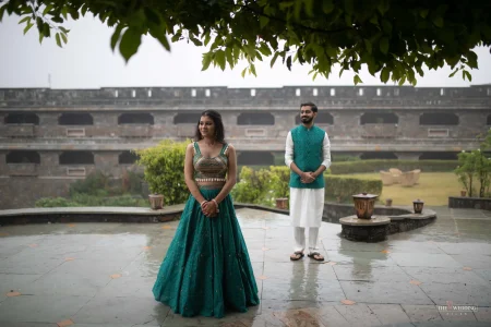 Couple posing separately at a historic Rajasthani fort with lush gardens, bride in teal embroidered lehenga and groom in teal waistcoat with white kurta, during a pre-wedding photoshoot at a heritage venue in Udaipur