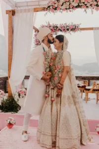 Bride in sequined ivory lehenga and groom in white sherwani sharing a close romantic moment after garland exchange during their rooftop wedding ceremony in Udaipur with floral mandap and misty hills in background