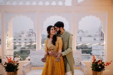 Couple in a close embrace on a palace rooftop during mehendi ceremony with Udaipur city skyline in background at dusk, bride in mustard yellow anarkali with intricate mehndi designs and groom in sage green sherwani
