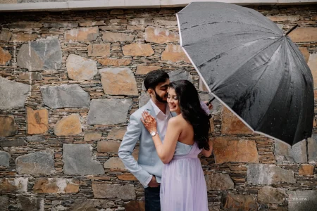 Couple sharing an intimate moment under a black umbrella against a rustic stone wall during a monsoon pre-wedding photoshoot in Udaipur, bride in lavender strapless flowy gown and groom in light blue blazer