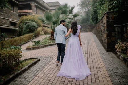 Couple walking together on a brick garden pathway at a luxury resort in Udaipur, bride in lavender flowy gown and groom in light blue blazer with dark jeans, captured from behind during a misty pre-wedding photoshoot