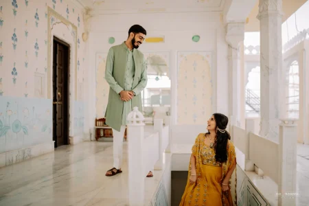 Couple sharing a candid laughing moment inside a painted royal haveli in Udaipur, groom in mint green sherwani standing on an upper step looking down at bride in mustard yellow ethnic wear seated below, with floral wall murals in background