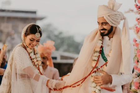 Bride in cream embroidered lehenga holding a garland while smiling at groom in ivory sherwani with cream turban during varmala exchange at an outdoor destination wedding ceremony in Udaipur Rajasthan