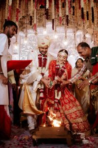 Bride in red embroidered lehenga and groom in white sherwani performing pheras around sacred fire under hanging garland backdrop at palace wedding ceremony