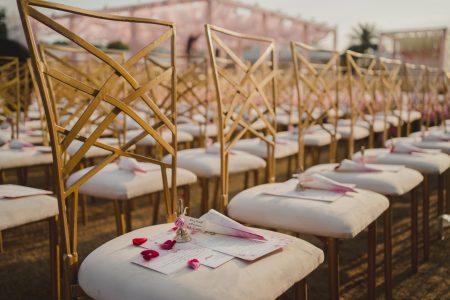 Rows of gold lattice back chairs with personalised wedding program scrolls and rose petals at outdoor wedding ceremony, The Leela Palace Jaipur destination wedding planned by Desert Pearl Weddings