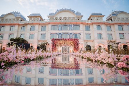 Wide shot of The Leela Palace Jaipur facade with pink floral arch mandap and stage reflected in foreground pool at golden hour, luxury destination wedding planned by Desert Pearl Weddings