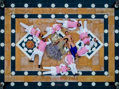 Stunning aerial drone photograph of bride in a navy blue gold embroidered lehenga and groom in a peach floral sherwani lying at the centre of an ornate geometric star-patterned heritage palace floor surrounded by eight groomsmen in matching pink bandhgalas and white churidars arranged in a radial pattern, at a luxury destination wedding in Udaipur Rajasthan