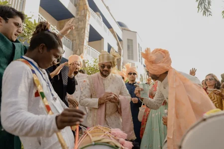 Groom in cream and blush pink embroidered sherwani with gold checkered turban and sunglasses dancing near a dhol player during baraat procession, surrounded by family and friends in vibrant ethnic wear at a luxury boutique destination wedding venue in Rajasthan