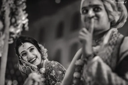 Black and white candid close-up of bride wearing a large maang tikka diamond headpiece floral jhoomar hair accessory layered pearl and polki necklace and embroidered bridal outfit laughing with genuine joy while looking up at groom in a safa turban who holds his finger to his lips in a playful shushing gesture in the soft-focus foreground during a wedding ceremony with blurred floral mandap decorations visible beside them at a luxury destination wedding in Rajasthan