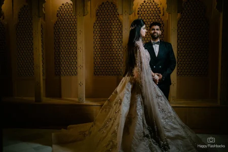 Bride in a stunning silver and blush ball gown lehenga with a long heavily embroidered floor-trailing skirt and feathered cape shown in side profile as groom in dark pinstripe suit with bow tie stands behind her gazing at her against a richly lit traditional Rajasthani geometric jali lattice stone wall at a luxury wedding in Rajasthan