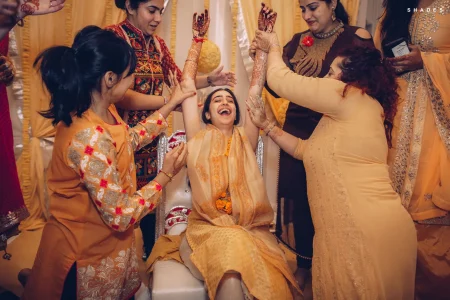 Bride in a gold and yellow tissue outfit with orange marigold garland and turmeric on her face throwing both arms up in pure joy with eyes closed and mouth open in laughter as five female relatives and friends in orange mustard and brown traditional Indian outfits hold up her mehndi-covered hands from all sides during a lively haldi ceremony with yellow draped fabric backdrop at a destination wedding in Rajasthan