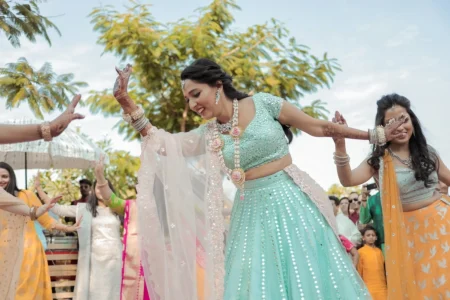 Bride in aqua teal mirror work sequin lehenga laughing and dancing joyfully with arms outstretched and dupatta flowing, mehndi designs visible on her raised hands, with female wedding guests in colorful ethnic wear celebrating in the background at an outdoor destination wedding in India