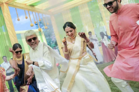 Bride in traditional cream Kerala kasavu saree with gold temple jewellery and mehndi dancing joyfully with an older family member in a white shirt and the groom in a pink kurta, all laughing together under a yellow and lime green outdoor canopy at a South Indian destination wedding celebration in India