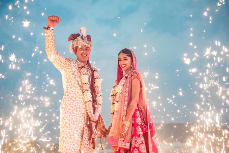 Bride in red embroidered lehenga and groom in cream gold sherwani with red turban celebrating joyfully together surrounded by dramatic ground sparklers against a blue sky after their wedding ceremony at a luxury destination wedding in Udaipur, groom pumping his fist in the air in a candid moment of joy